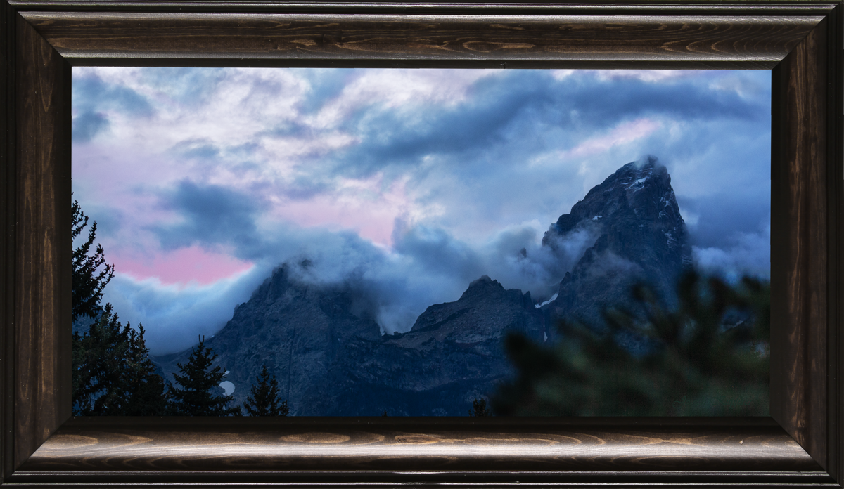 A framed landscape art print depicting a majestic mountain peak shrouded in dramatic clouds with a soft pink glow on the horizon, enclosed in a dark brown wooden frame.